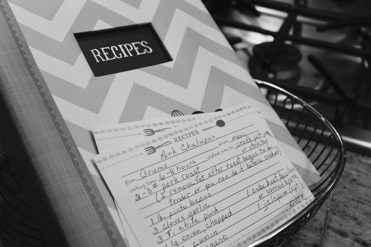 heros-img Monochrome image of a recipe book and handwritten notes in a kitchen setting.
